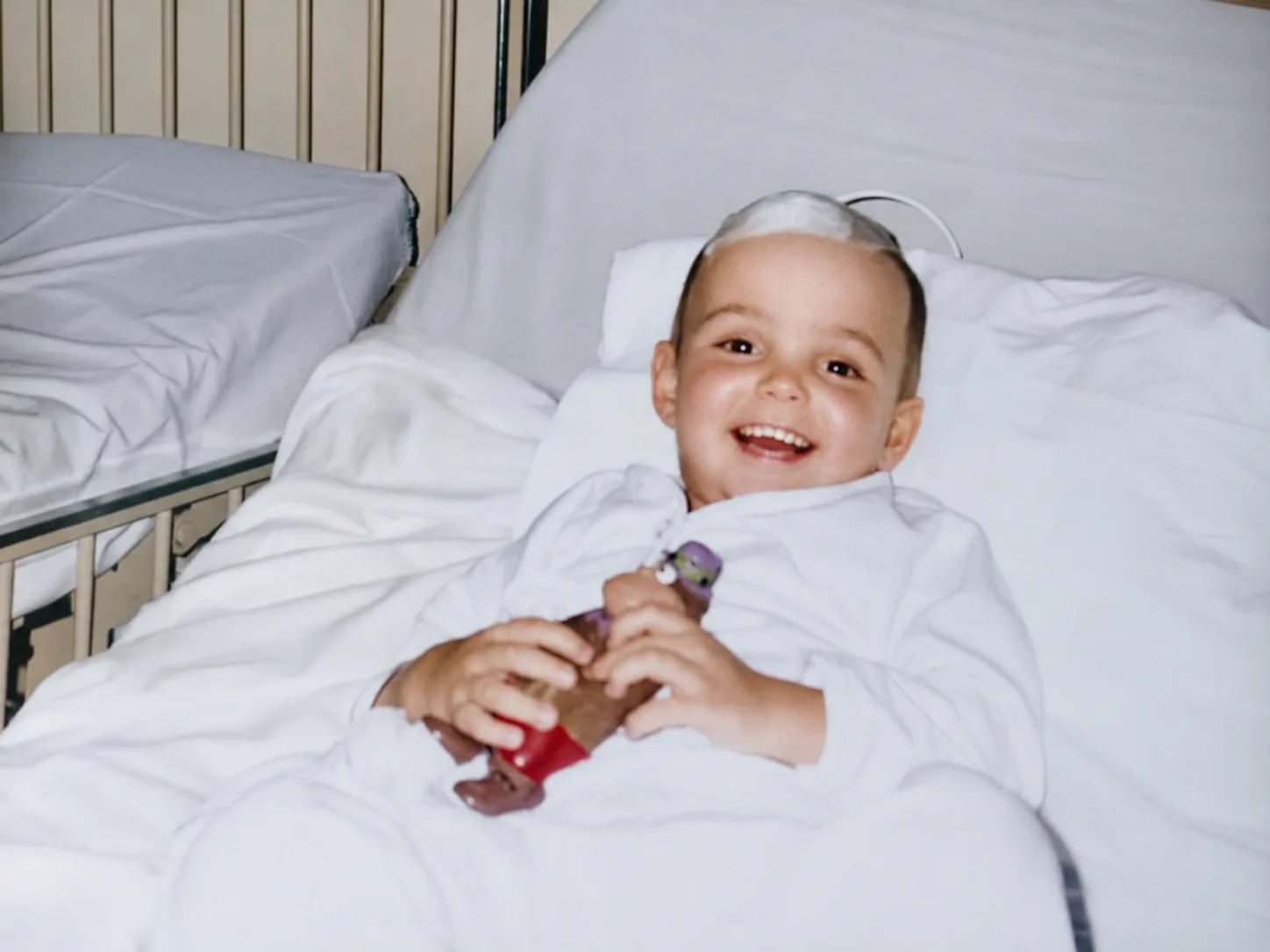 Dr. João Guttierrez ainda criança, sorrindo em uma cama de hospital pouco depois de uma cirurgia, com um curativo na cabeça.